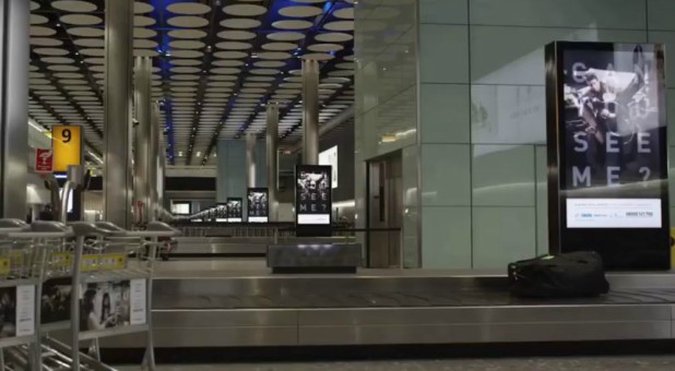 Can You See Me? posters line an airport baggage claim.